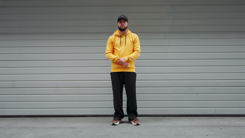 Young man captured mid-dance move in yellow hoodie and black cap stands against gray industrial background. Pose urban attire reflect energy and vibe of modern street dance culture. Freedom music styl