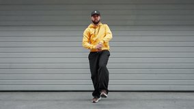 Young man captured mid-dance move in yellow hoodie and black cap stands against gray industrial background. Pose urban attire reflect energy and vibe of modern street dance culture. Freedom music styl - Powered by Shutterstock - Get 15% off with code: PIKWIZARD15