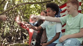 Four male and female multi-ethnic school children wearing casual clothes, sitting in front of USA flag in forest reaching hands to light sparklers on Independence Day - Powered by Shutterstock - Get 15% off with code: PIKWIZARD15