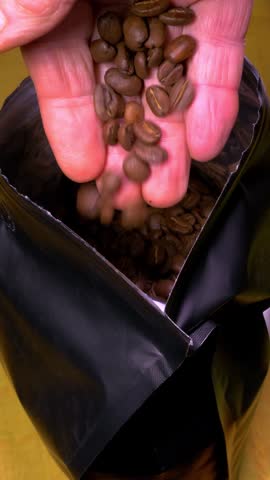 Vertical video - Closeup of a man’s hand dipping into a newly opened pack of fresh brown roasted coffee beans, then lifting a handful out to take a look, before dropping them back in.