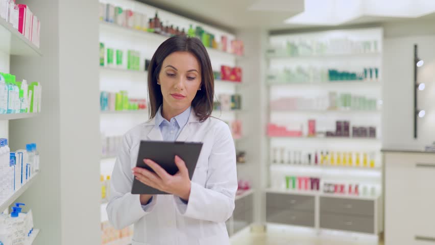 Female pharmacist standing near racks with cosmetics. Woman druggist conducting audit of new supply of medicine. Woman pharmacist holding tablet. Female druggist wearing white gown in drugstore.