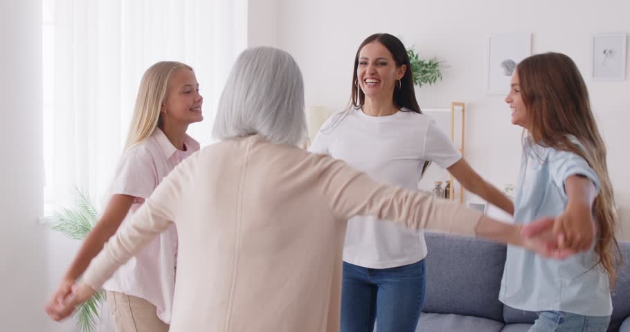 Happy multi generation family jumping high and dancing in round dance in living room. Four women daughters, mother and grandmother having fun, holding hands, enjoying leisure time together at home.