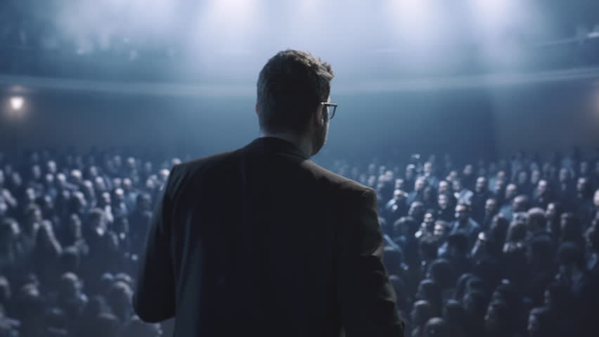 Silhouetted Speaker On Stage Addressing Large Crowd In Dark Auditorium Leadership Influence Power