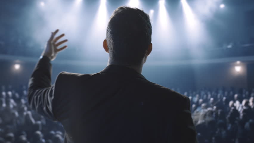 Silhouetted Speaker Addresses Large Crowd In Dark Arena Success Leadership Power