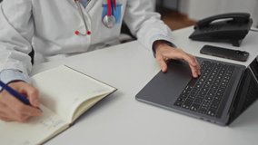 Doctor writing notes in notebook while using laptop in hospital office showing hands on keyboard and pen on paper indicating work in medical interior setting with focus on technology. - Powered by Shutterstock - Get 15% off with code: PIKWIZARD15