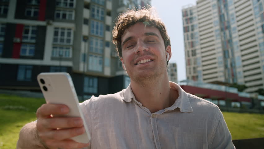 Caucasian man male guy sitting outside holding mobile phone smartphone technology device headphones listening music enjoyment relax playlist earphones businessman mp3 wireless song city gadget weekend