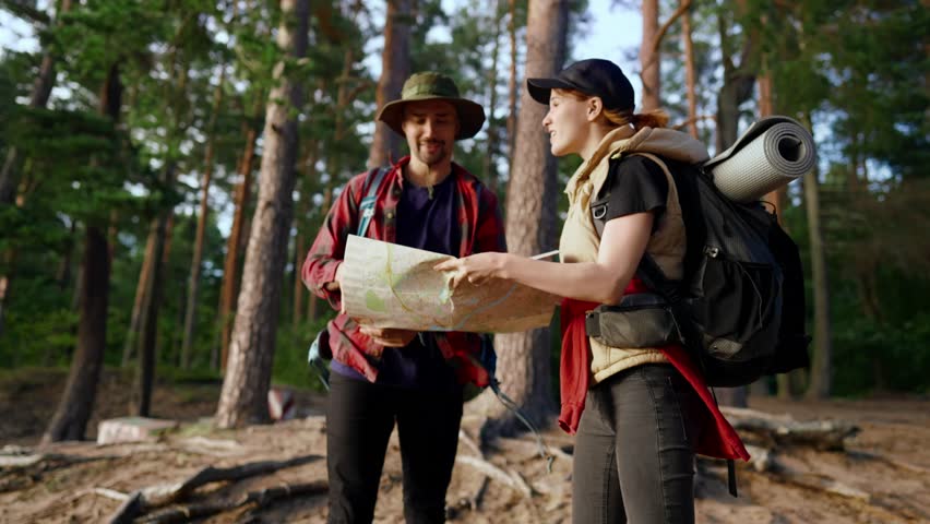 Backpackers lost in forest, scared man and woman looking for exit from woodland . Pair of adult tourists with backpacks holding paper map of area of national park, traveling in nature in summer day