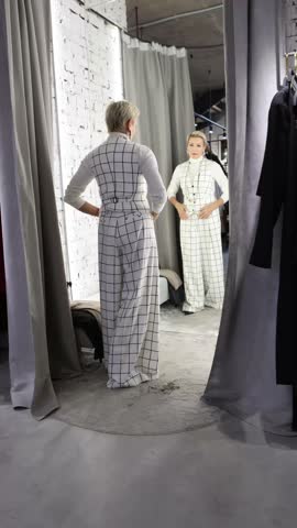 Stylish Woman Trying On and Selecting Fashionable Clothing in Fitting Room of Female Fashion Store, Featuring Chic Checkered White Suit While Looking in Mirror