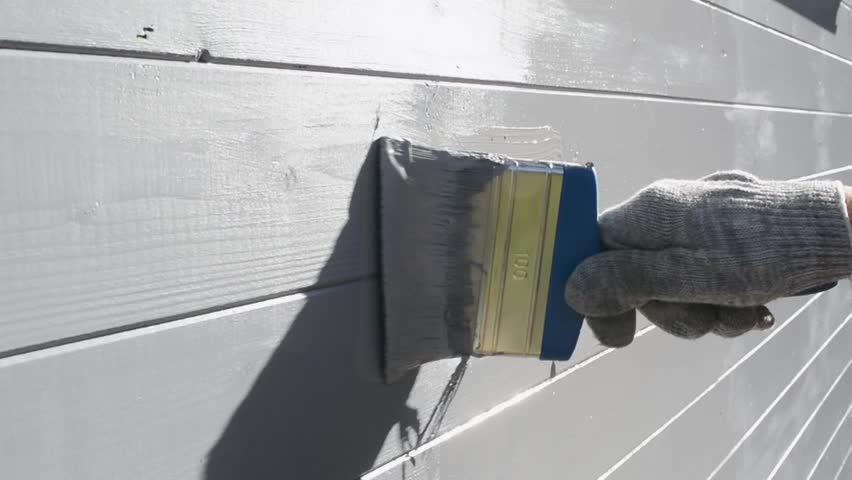 A brush applies gray paint to the wooden house walls, leaving a smooth and uniform coating. The process of protecting and painting the facade.