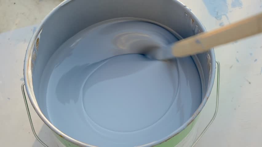 A close-up of gray paint being stirred smoothly in a paint can with a wooden stir stick. The paint swirls evenly as it is prepared for application in a DIY or home improvement project.