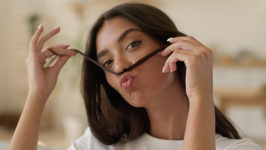 Happy smiling positive playful funny beautiful Indian Arabian woman girl female lady playing curl hair put like mustache grimace pretty at home indoors smooth hairstyle color hairdo shampoo cosmetic