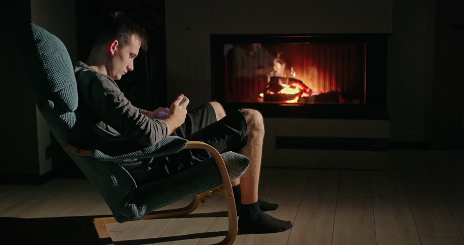 Young man scrolling on a smartphone while seated in a cozy armchair near a glowing fireplace, enjoying a warm and tranquil ambiance 
