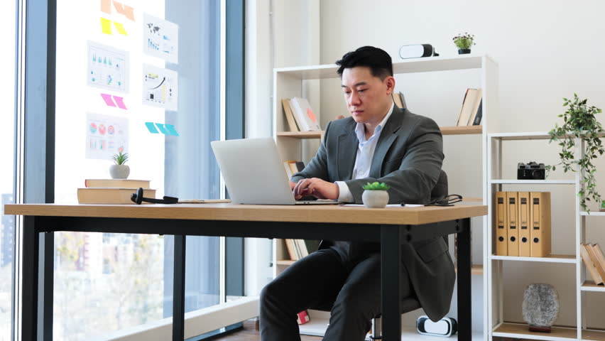 Asian businessman in modern office focused on laptop while writing report. Wearing formal attire, mid-aged male demonstrates concentration in professional environment. - Powered by Shutterstock - Get 15% off with code: PIKWIZARD15