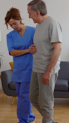 Vertical video of mid adult nursing home helping older man to move at domestic room. Medical female worker in medical uniform supporting senior adult man wheelchair.