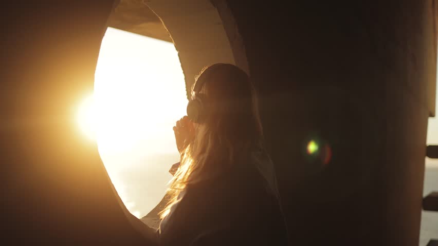 Atmospheric, cinematic video of a girl in headphones listening to music near a round window overlooking the sunset and the ocean, music lover, teenager, sun rays, golden hour, aesthetics, feelings