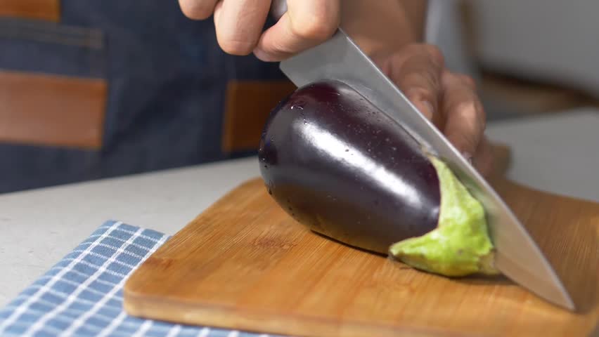 Slow Motion Side Perspective of a Chef Slicing an Eggplant on a Wooden Cutting Board with a Blue Plaid Cloth, Focusing on the Process of Crafting a Vegetable-Based Dish. Vegetarian Concept