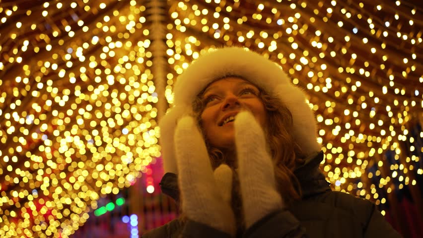 Young woman is smiling while looking up at christmas lights at a holiday market. She is wearing a white hat and gloves and a black coat