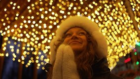 Young woman is smiling while looking up at christmas lights at a holiday market. She is wearing a white hat and gloves and a black coat - Powered by Shutterstock - Get 15% off with code: PIKWIZARD15