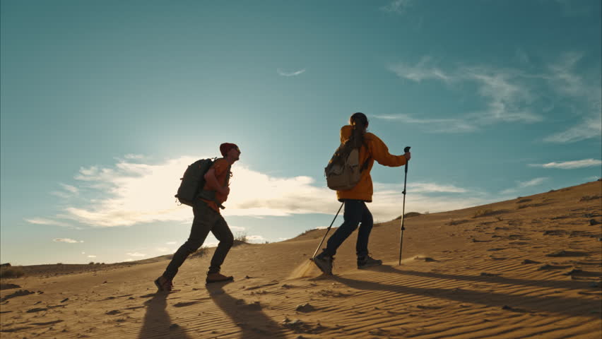 Couple of active hikers walks with hiking poles and backpacks at sunset desert dunes against blue sky