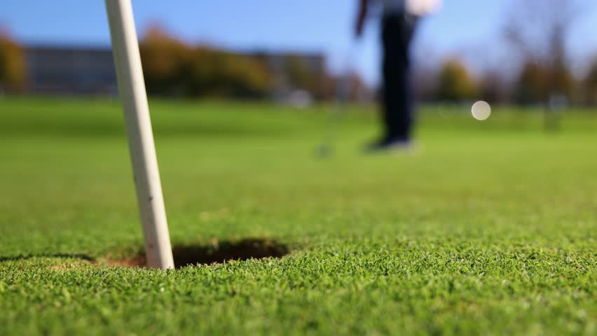 close-up shot at ground level captures a golf ball rolling into the hole after being hit by a senior golfer in the background. highlights the precision and focus of the golfer for a successful shot
