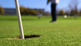 close-up shot at ground level captures a golf ball rolling into the hole after being hit by a senior golfer in the background. highlights the precision and focus of the golfer for a successful shot - Powered by Shutterstock - Get 15% off with code: PIKWIZARD15