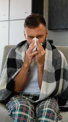 Sick Man Sneezing into Tissue, Covered in Blanket on Couch