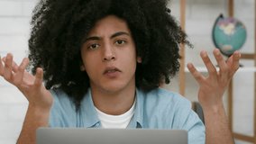 A young man with curly hair sits at a desk, looking confused and raising his hands in a questioning gesture while using his laptop. The setting is a cozy, well-lit indoor space. - Powered by Shutterstock - Get 15% off with code: PIKWIZARD15