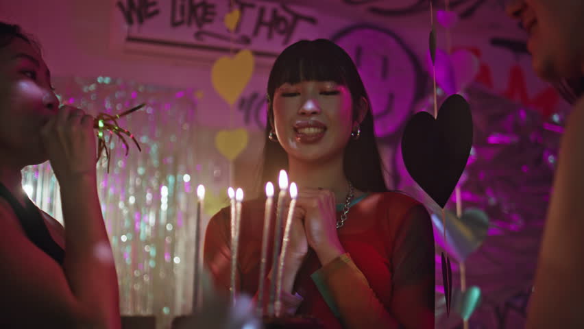Happy woman blowing candles on cake at bright nightclub closeup. Joyful asian girl celebrating birthday with friends in vivid club. Group smiling youngsters congratulating smiling female with event.