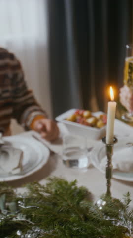 Vertical close-up shot of hands of friends clinking champagne glasses at elegantly decorated holiday table with candlelight and evergreen branches during Christmas celebration