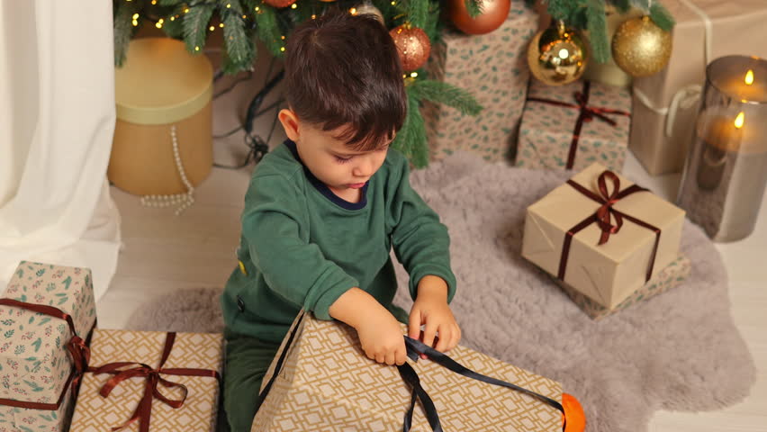 sleepy kid awaiting santa claus.adorable baby boy sitting next to christmas tree, unboxing a gift.kid raising up many boxes.child sitting in orange armchair with tangerine in hand. bokeh lights