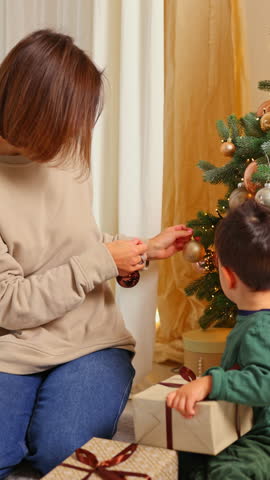 mother and son decorating christmas fir tree in cozy home interior.woman and boy kid putting globes balls on branch, playing covering eyes or unboxing gift.smiling female mom having fun with child.