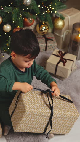 sleepy kid awaiting santa claus.adorable baby boy sitting next to christmas tree, unboxing a gift.kid raising up many boxes.child sitting in orange armchair with tangerine in hand. bokeh lights
