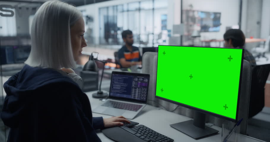 Caucasian Girl Working in a Modern Successful Company as a Software Engineer. Female Using a Desktop Computer with a Green Screen Mock Up Template on a Monitor. Additional Laptop Screen Showing Code
