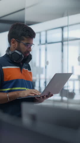 Vertical Screen: Portrait of an Indian Young Male Standing in Office with a Laptop Computer, Using Device to Monitor the Development of a Software Project. Manager Working in Modern IT Company Office