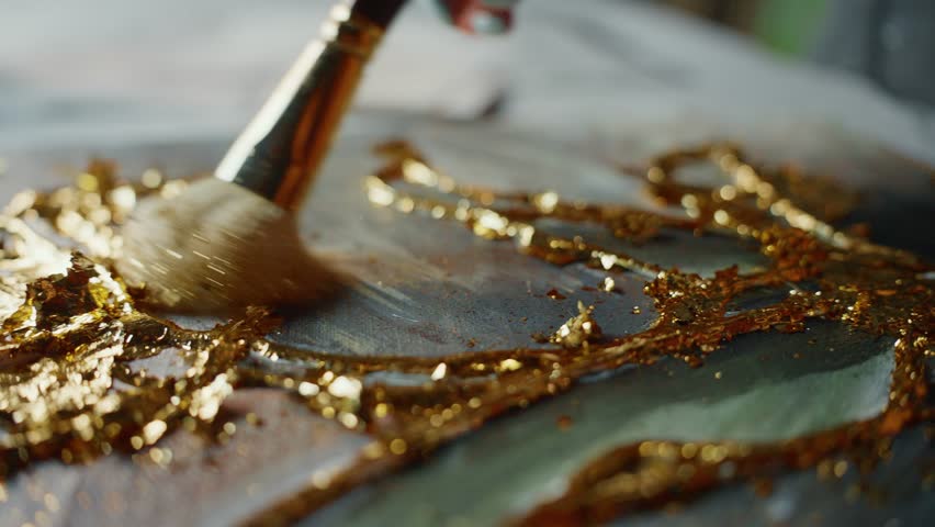 A close-up shot of an artist applying gold leaf to a resin artwork using a brush. The artist carefully applies the gold leaf to the surface, creating a shimmering, luxurious effect.