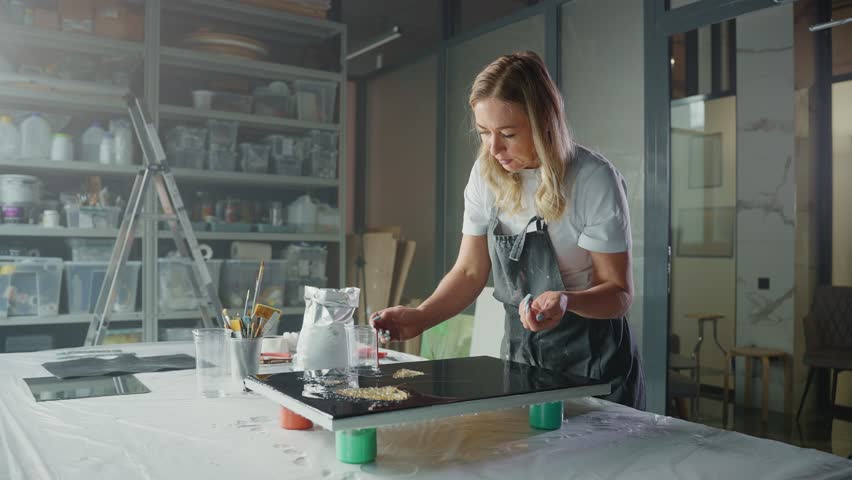 A woman in an apron works on an abstract artwork in her home studio. She is using gold resin and a tool to create a unique design on the canvas.