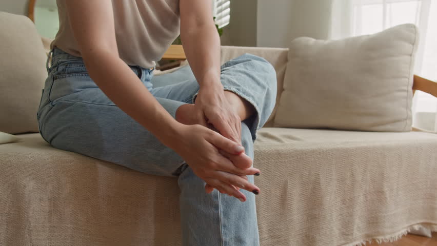 Cropped shot of anonymous woman in jeans moisturizing her feet, toes and heels with cream while sitting on couch