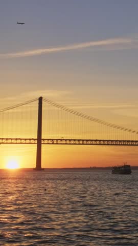 View of 25 de Abril Bridge famous tourist landmark of Lisbon connecting Lisboa and Almada over Tagus river with boat silhouette on sunset and flying plane. Lisbon, Portugal. Camera pan