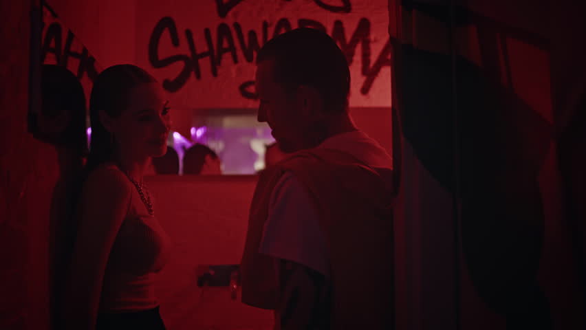 Young couple flirting on party at graffiti wall nightclub backdrop closeup. Red ambient light creating intimate atmosphere for pair romantic interaction. Beautiful woman talking to cool man in club.