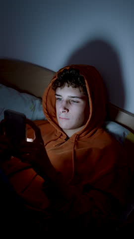 4k medium handheld vertical shot of teenager using smartphone lying in bed late at night, playing, watching videos, and scrolling the screen. Youth and Children's screen addiction.