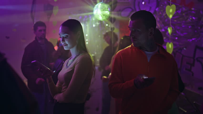 Couple with smartphones standing club ignoring each other under disco ball closeup. Man looking at woman using cellphone in vibrant nightclub. Two clubbers with phones meeting on vibrant dance floor.