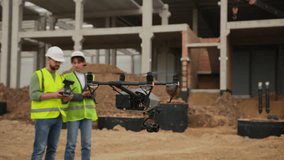 Two builders in bright yellow vests using drone on construction site. Gray and black drone with camera in foreground on blurred background. Outdoors - Powered by Shutterstock - Get 15% off with code: PIKWIZARD15