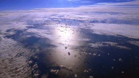 A high altitude aerial view over clouds on a sunny day, taken from the cabin of a passenger aircraft. The camera is stationary, tilted down looking out the port side window at the ocean and clouds. - Powered by Shutterstock - Get 15% off with code: PIKWIZARD15
