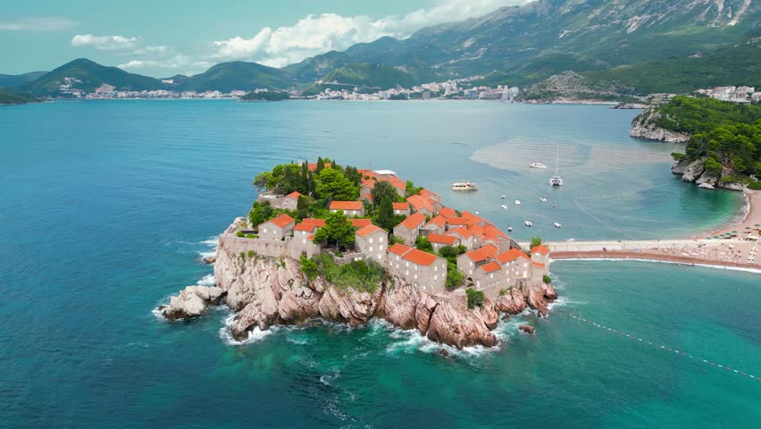 Sveti Stefan Island - Montenegro. Waves breaking on the rock. Famous and luxury travel destination. Boats in the bay. Turquoise water. Drone circling around the island
