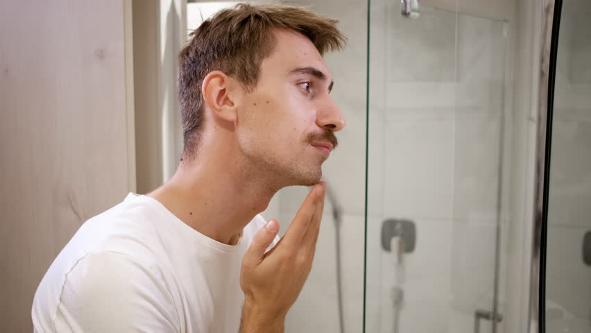 Young man with mustache checking out his skin in the bathroom mirror.