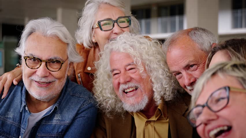 Cheerful group of senior citizens people laughing and enjoying video call embracing together. Retired generation community friends having fun gathered on terrace waving happily at camera