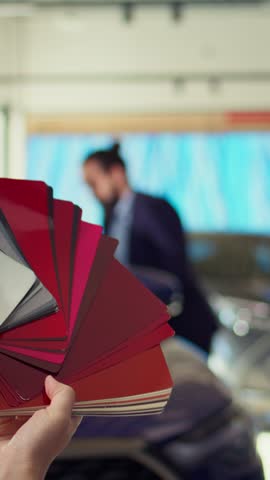 Vertical video Salesperson in auto showroom discussing with man, offering color swatch samples choice. Agent providing client with color wheel selection, picking hue for car paint, camera B close up