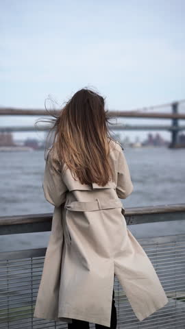A woman in a stylish trench coat gazes at a distant city bridge by the shimmering water in a calm urban setting. Stylish woman wearing trench coat stands by water gazing beautiful city bridge