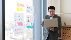 Asian businessman in professional suit analyzing charts and graphs during video call. Standing by panoramic window with laptop, pointing at data visuals. Modern office setting with urban view. - Powered by Shutterstock - Get 15% off with code: PIKWIZARD15