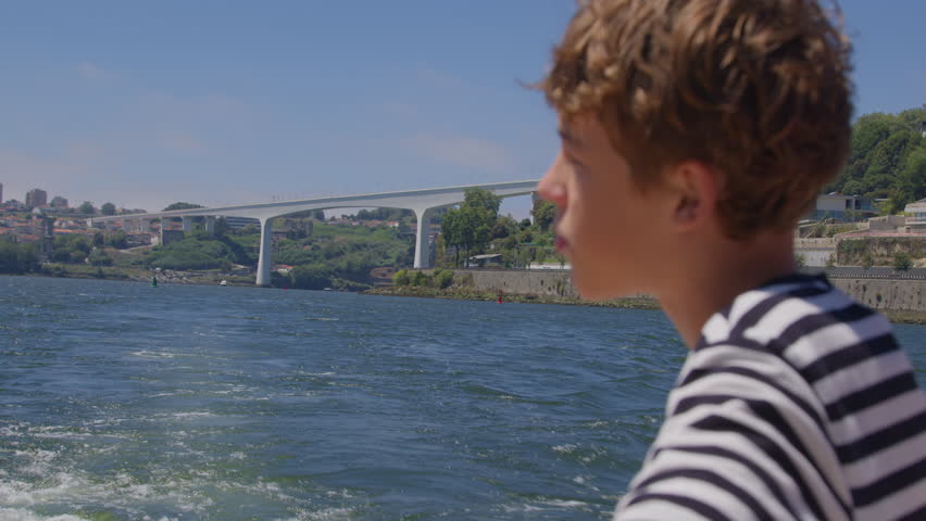 Young Caucasian boy gazing at a white bridge stretching over the river from the boat, with a city landscape and green hills in the background